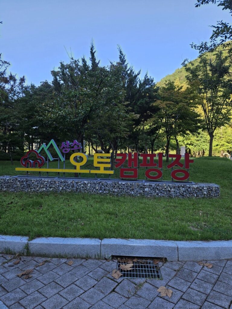 파란 하늘과 푸른 나무들 아래, '상소 오토캠핑장'이라는 글자가 다채로운 색상으로 쓰인 입구 표지판이 선명하게 보인다. 주변에는 초록색 잔디와 포장된 길이 보인다.