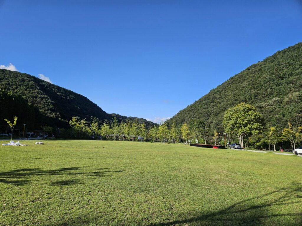 대전 상소동 산림욕장의 넓게 펼쳐진 푸른 잔디밭과 울창한 숲으로 둘러싸인 산, 맑고 푸른 하늘이 조화를 이루는 평화로운 풍경