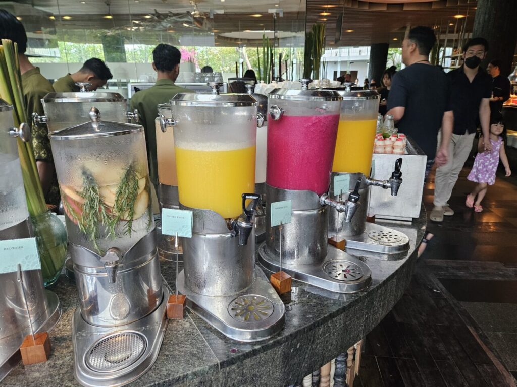 아야나 에스테이트 조식 뷔페의 음료 코너입니다. 다양한 종류의 신선한 과일 주스가 대형 디스펜서에 담겨 있으며, 과일과 허브를 넣은 얼음물이 준비되어 있습니다.