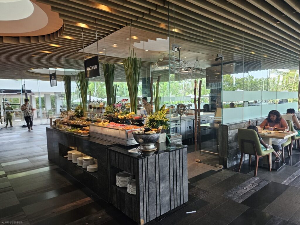 발리 아야나 에스테이트의 현대적이고 개방감 있는 뷔페 레스토랑 전경입니다. 다양한 음식 코너가 마련되어 있으며, 창밖으로 푸른 정원이 보이는 곳에서 손님들이 식사하고 있습니다.