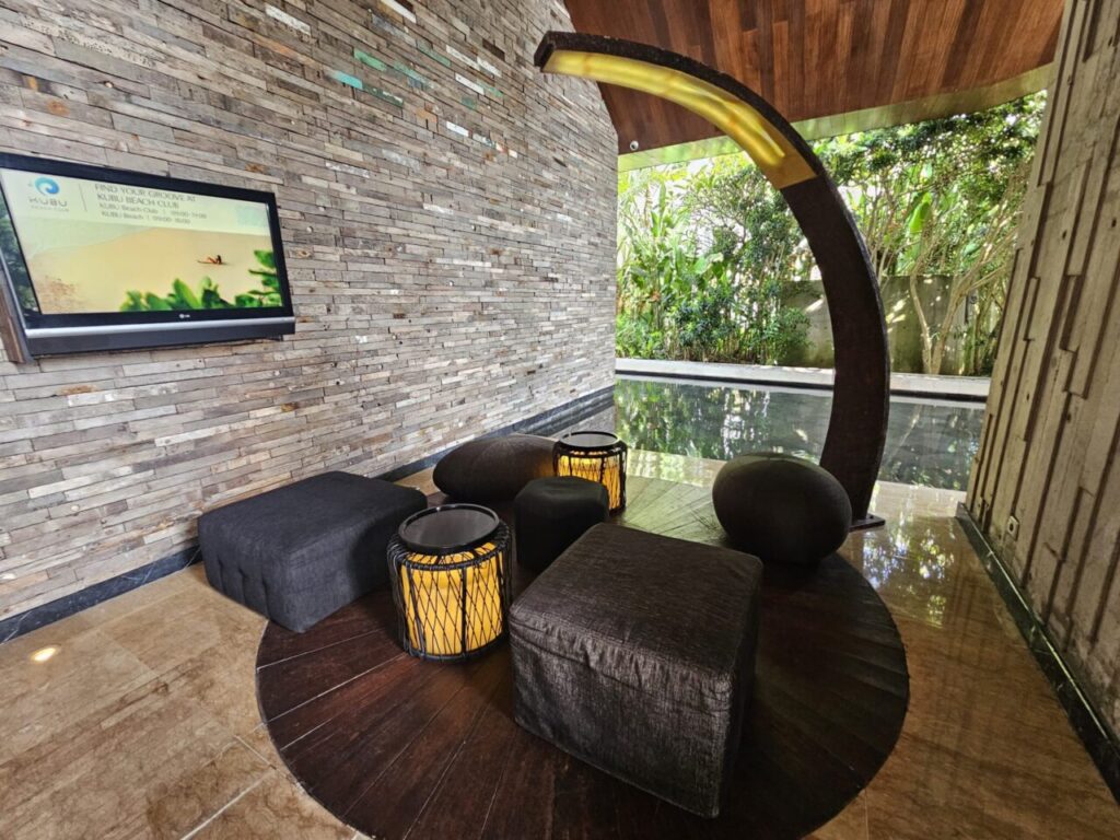 아야나 에스테이트의 세련된 실내 휴게 공간입니다. 현대적인 의자와 쿠션, 독특한 곡선형 스탠드 조명, 벽걸이 TV가 어우러져 편안한 분위기를 연출합니다.