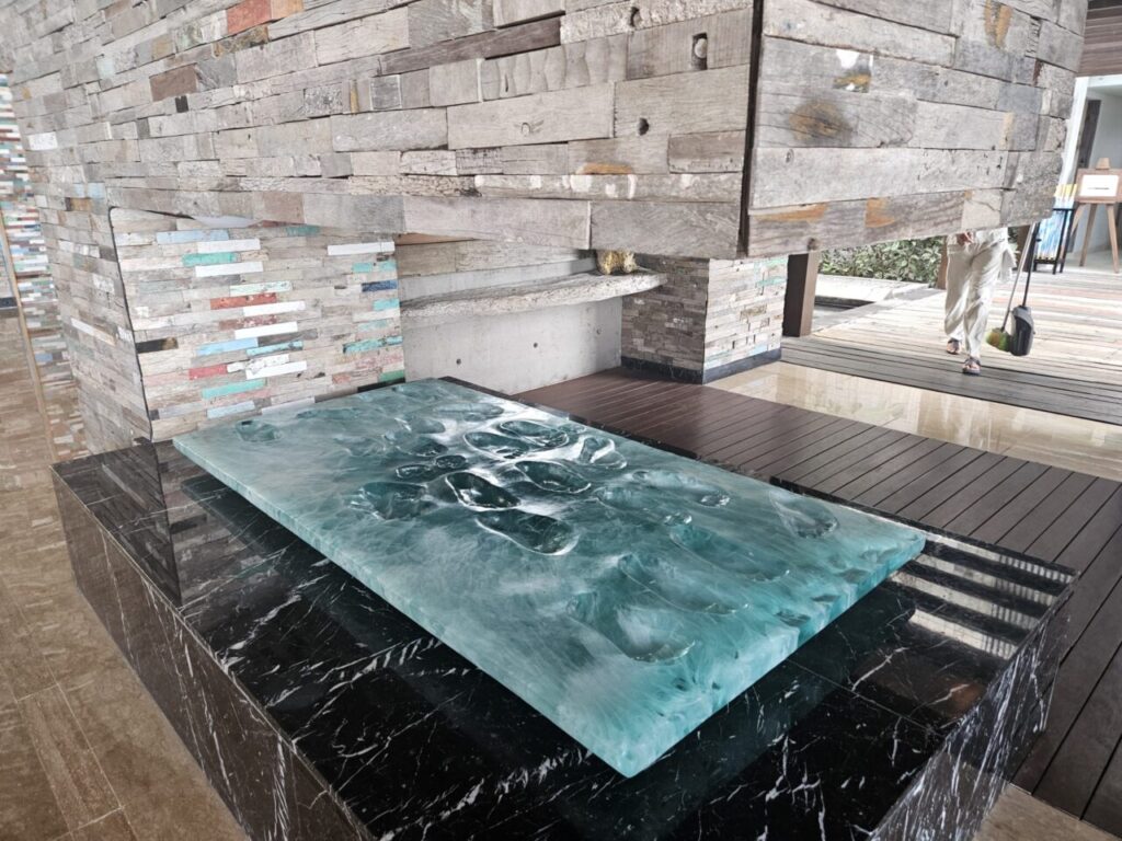 아야나 에스테이트의 현대적이고 예술적인 리셉션 또는 라운지 카운터입니다. 질감이 느껴지는 나무 벽과 물결무늬의 투명한 초록색 상판이 인상적입니다.