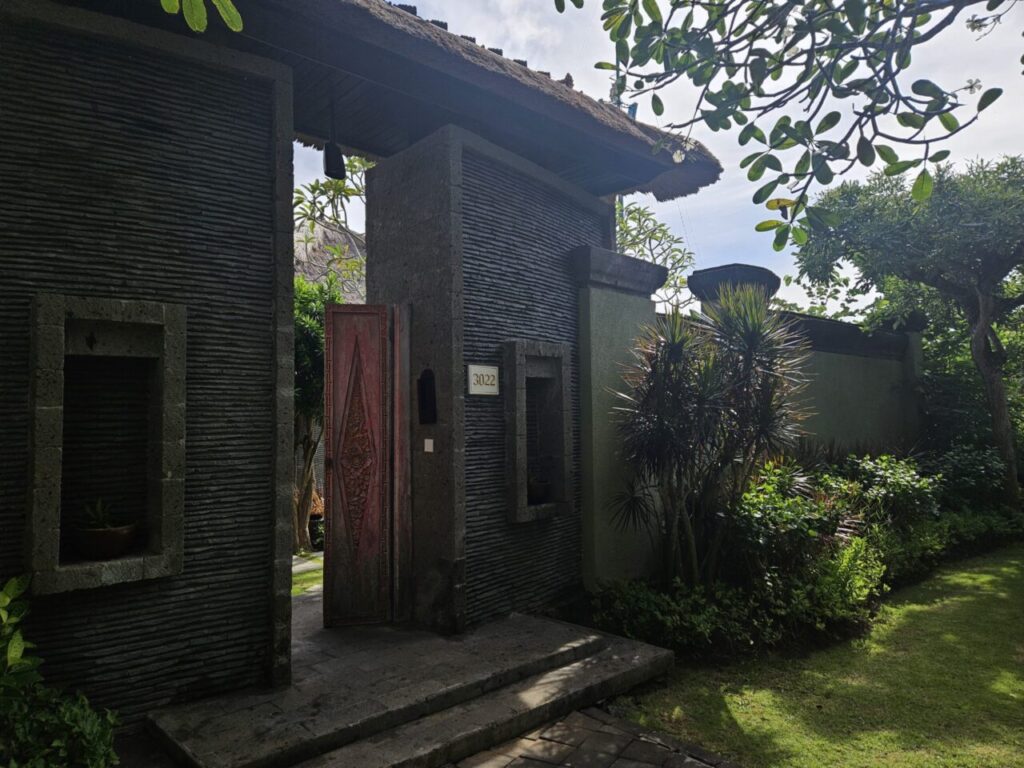 발리 아야나 에스테이트 빌라의 입구입니다. 돌담에 새겨진 독특한 붉은색 조각 나무문이 인상적이며, 주변은 푸른 식물들로 가득합니다.