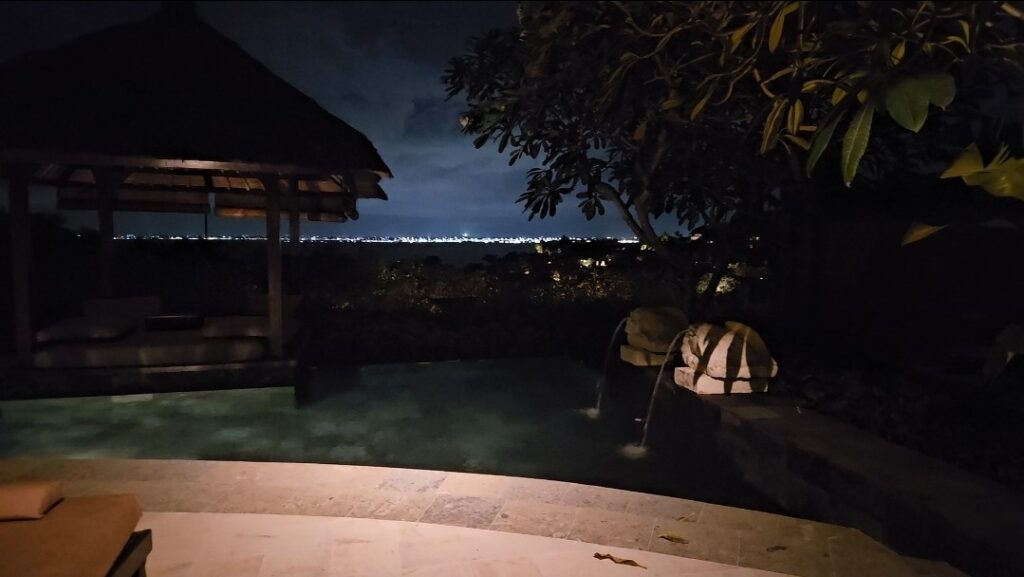 밤에 촬영된 아야나 에스테이트 빌라의 개인 수영장 공간입니다. 은은한 조명 아래 가제보와 물 조형물이 보이며, 멀리 해안선을 따라 도시의 불빛이 반짝입니다.