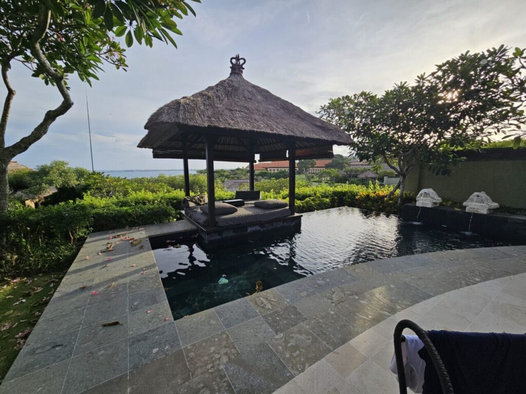 발리 아야나 에스테이트 빌라의 개인 수영장 공간입니다. 초가지붕의 가제보와 일광욕 의자들이 놓여 있고, 멀리 바다가 보이는 아름다운 풍경을 자랑합니다.
