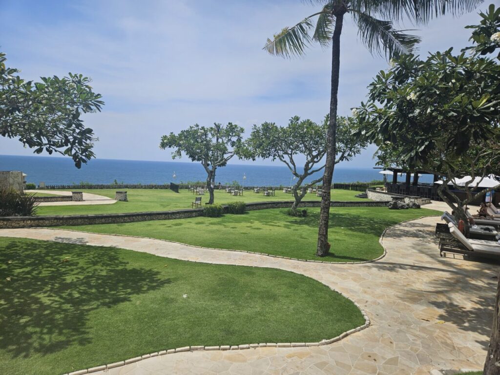 발리 아야나 리조트에서 바라본 넓은 잔디밭과 푸른 바다 전경. 멀리 바다를 배경으로 테이블과 의자들이 놓여 있다.