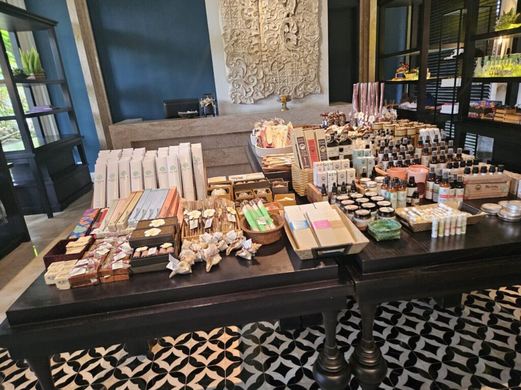 발리 아야나 리조트 내 상점의 전경으로, 다양한 종류의 향수, 오일, 수공예품 등이 진열된 넓은 테이블과 선반이 보인다.