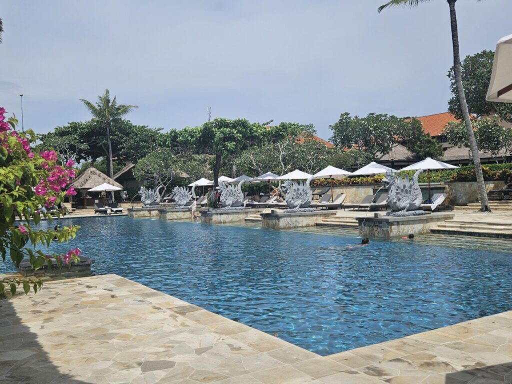 발리 아야나 리조트의 넓은 수영장. 푸른 물과 함께 독특한 석상들이 놓여 있고, 주변에는 편안한 선베드와 파라솔이 배치되어 있다.