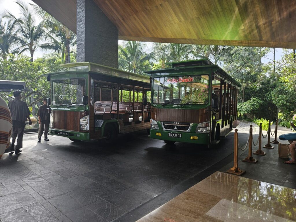 발리 아야나 리조트의 트램 정류장에 주차된 두 대의 초록색과 갈색 트램 차량. 나무 벤치가 설치되어 있고 직원들이 대기하고 있다.