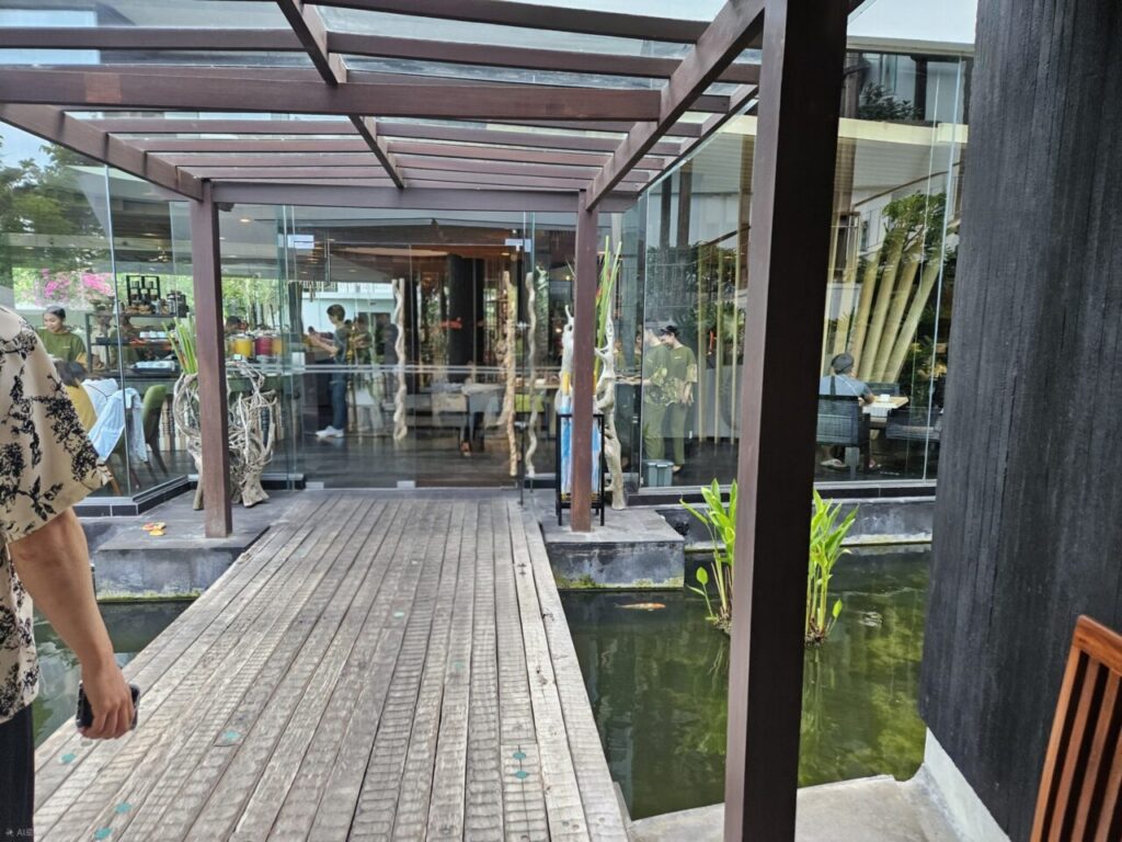 푸른 식물과 물이 있는 연못 위를 가로지르는 나무 보행로입니다. 길 끝에는 현대적인 유리벽 건물이 보이며, 실내에는 직원들이 분주하게 움직이고 있습니다.