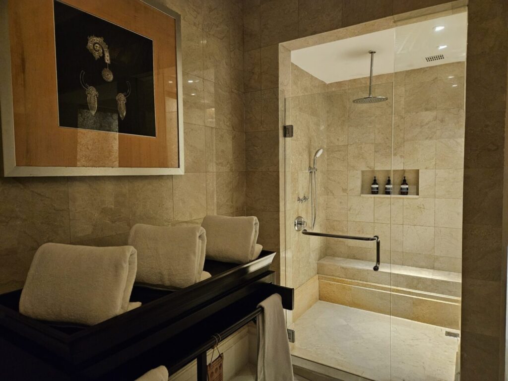 고급스러운 욕실 내부에 넓은 유리 칸막이 샤워 부스입니다. 천장형 레인 샤워기와 벽 선반에 놓인 목욕 용품들이 보이며, 편안하게 앉을 수 있는 벤치도 있습니다.