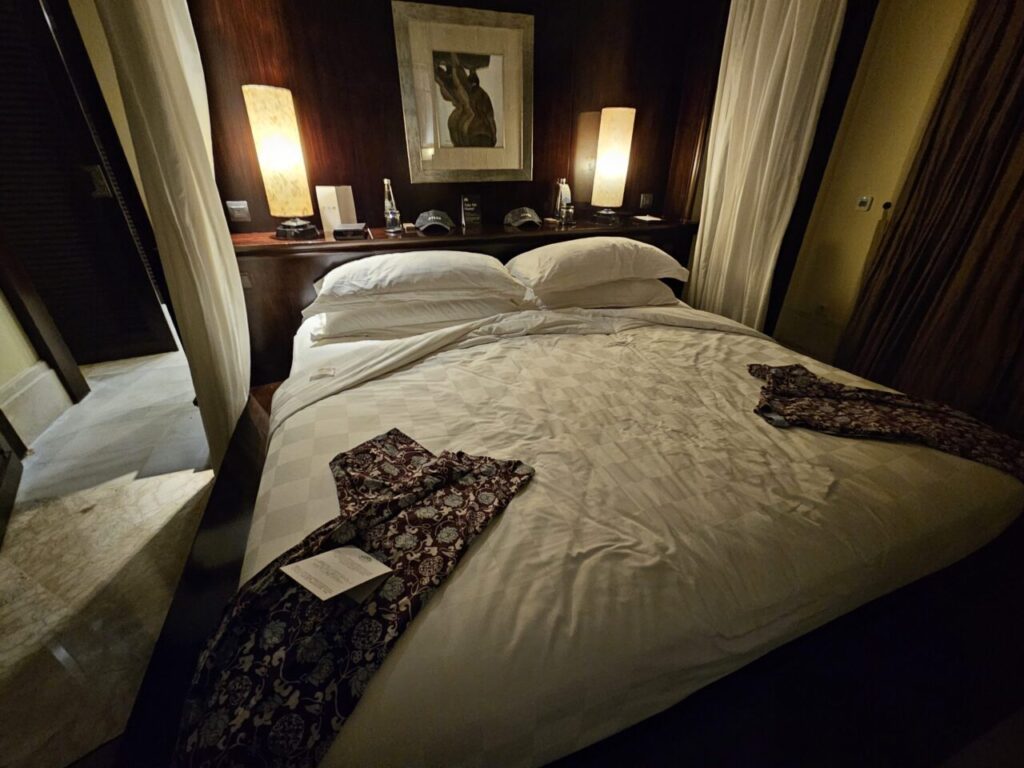 아야나 빌라 침실의 깔끔하게 정돈된 침대입니다. 하얀색 침구와 장식용 베개가 놓여 있고, 전통적인 인도네시아 바틱 문양의 사롱이 침대 위에 펼쳐져 있습니다.