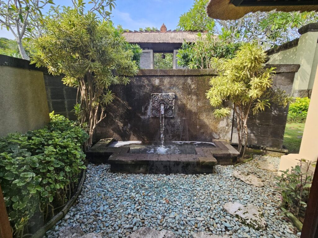 빌라 입구에 위치한 작은 돌 분수입니다. 조각된 돌 벽에서 물이 흘러내려 자갈이 깔린 바닥으로 떨어지고, 주변은 싱그러운 녹색 식물로 둘러싸여 있습니다.