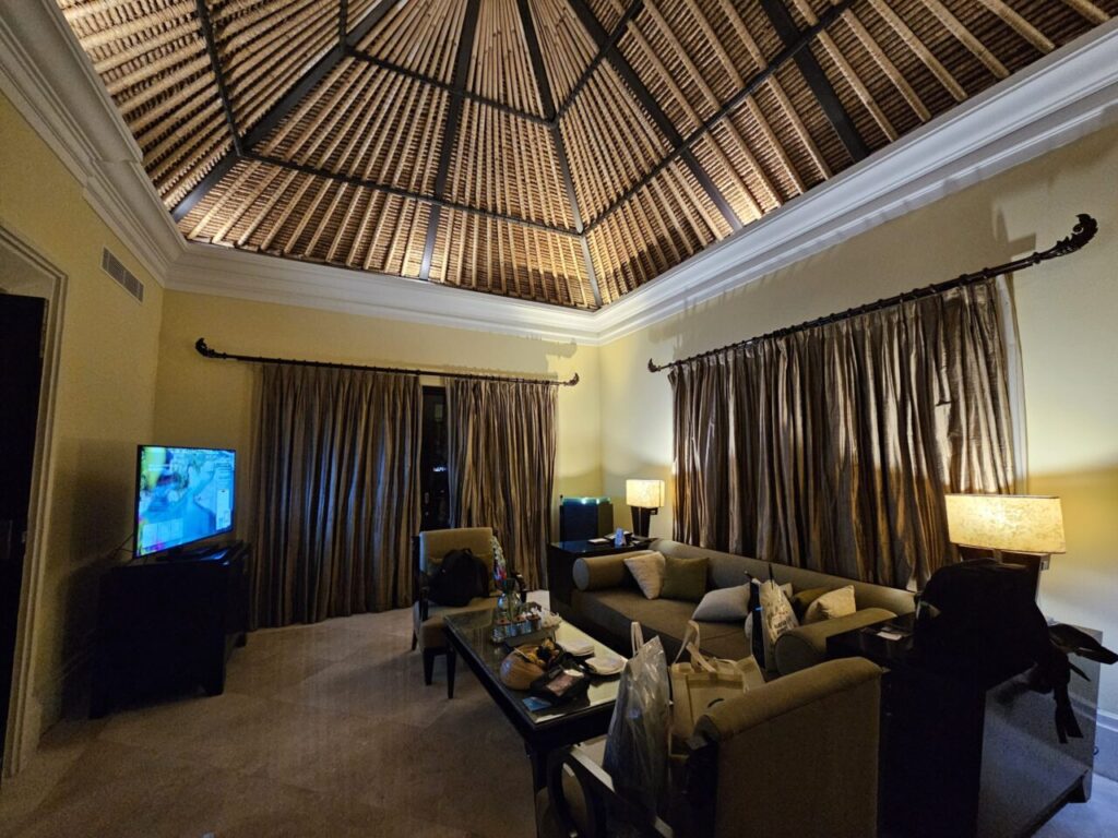 아야나 빌라의 넓고 고급스러운 거실 전경입니다. 편안한 소파와 커피 테이블, 그리고 독특한 직조 패턴의 높은 천장이 특징입니다.