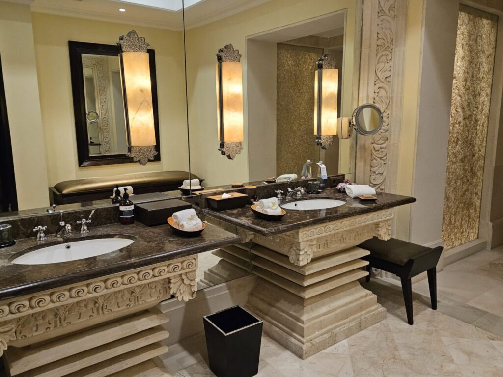 아야나 빌라 욕실의 고급스러운 세면대 공간입니다. 두 개의 세면볼과 화려하게 장식된 거울, 우아한 조명이 조화를 이루며 럭셔리한 느낌을 줍니다.