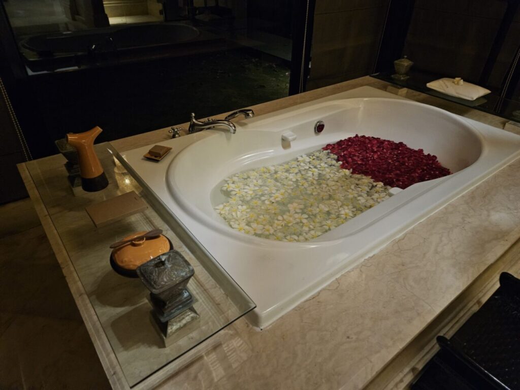 물과 붉은색, 흰색 플루메리아 꽃잎으로 가득 채워진 넓은 욕조입니다. 로맨틱하고 편안한 분위기를 연출하며, 옆에는 욕조 용품들이 놓여 있습니다.