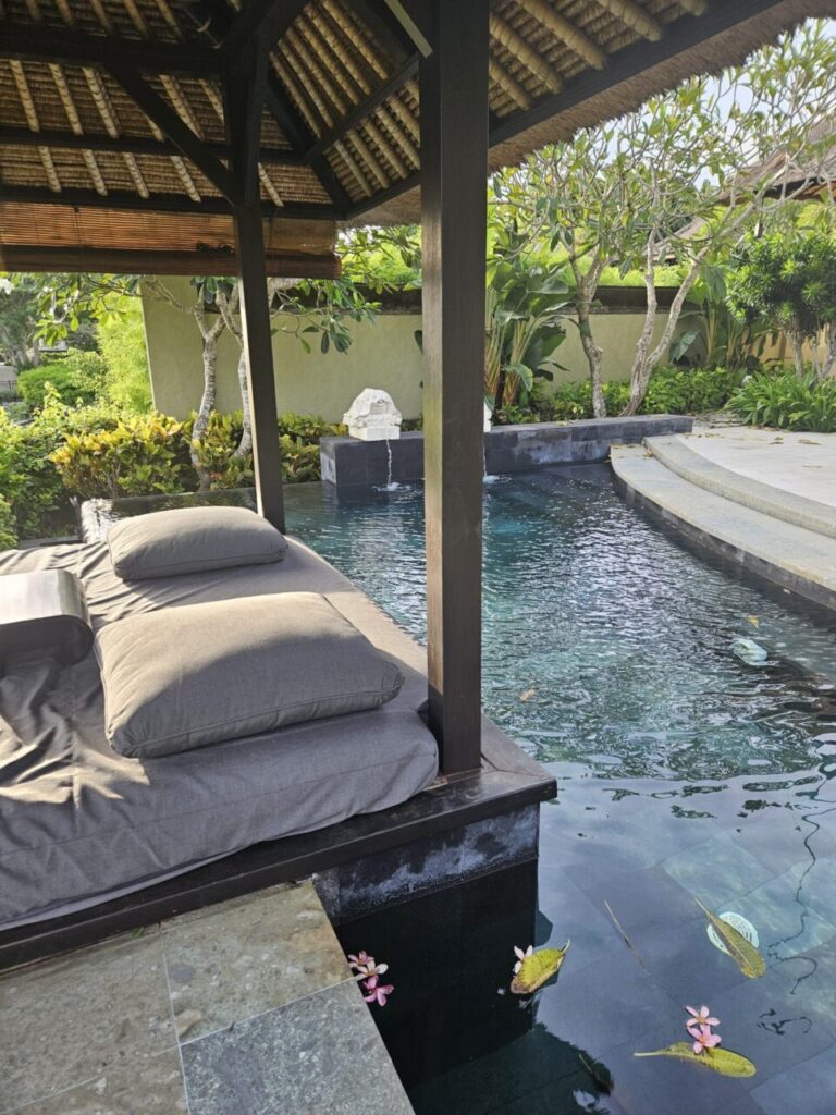 아야나 빌라의 개인 수영장 옆에 있는 아늑한 카바나입니다. 물 위에는 플루메리아 꽃잎들이 떠 있고, 편안한 쿠션이 놓여 있어 휴식을 취하기 좋습니다.