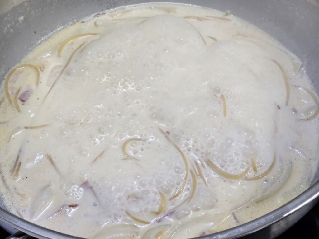 냄비 안에 베이컨 조각들이 섞인 크림 소스와 스파게티 면이 끓고 있는 모습