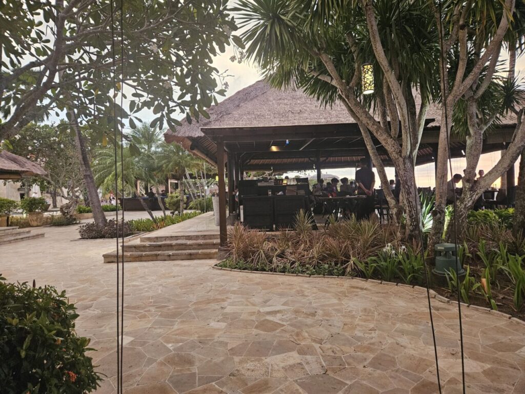 발리 아야나 리조트의 아름다운 야외 레스토랑 전경. 초가지붕 아래에서 식사하는 손님들과 열대 나무들, 돌 포장길이 어우러져 이국적인 분위기를 연출한다.