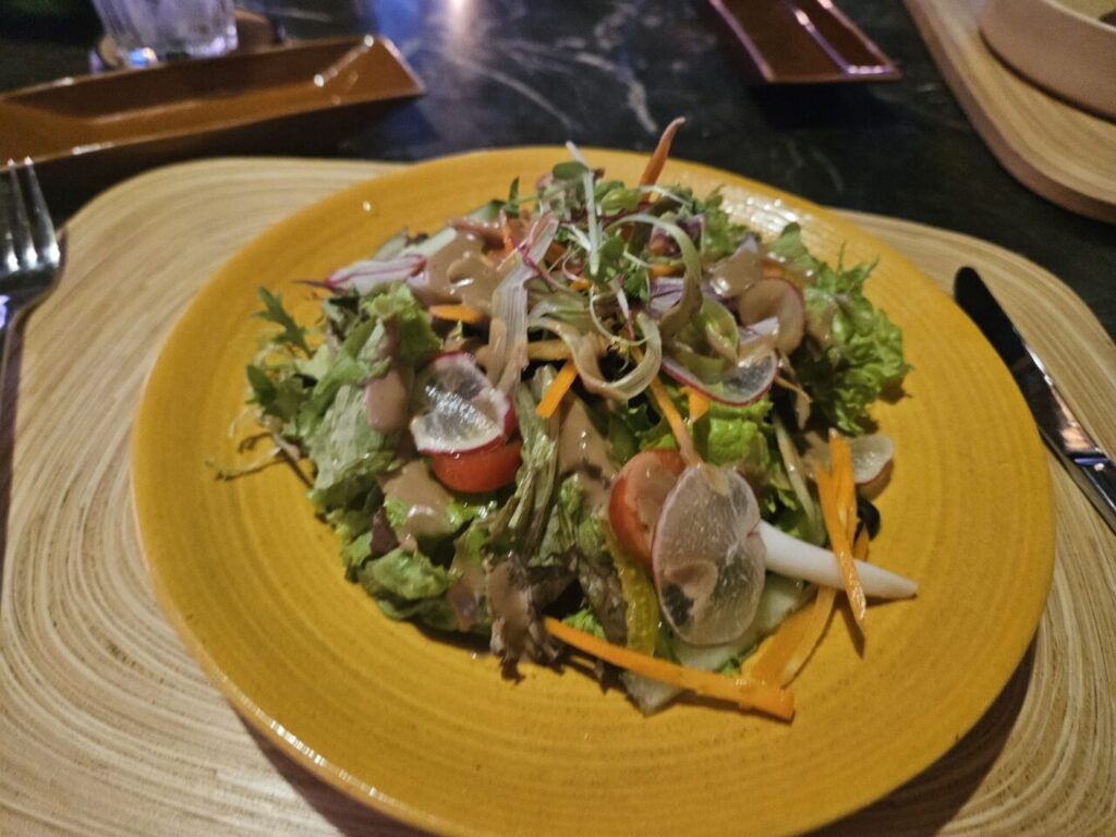 발리 아야나 리조트의 한 식당에서 찍은 사진으로, 대나무 매트 위에 놓인 노란색 접시에 신선한 채소와 크리미한 드레싱이 어우러진 샐러드가 담겨 있다. 식사용 포크와 나이프가 옆에 보인다.