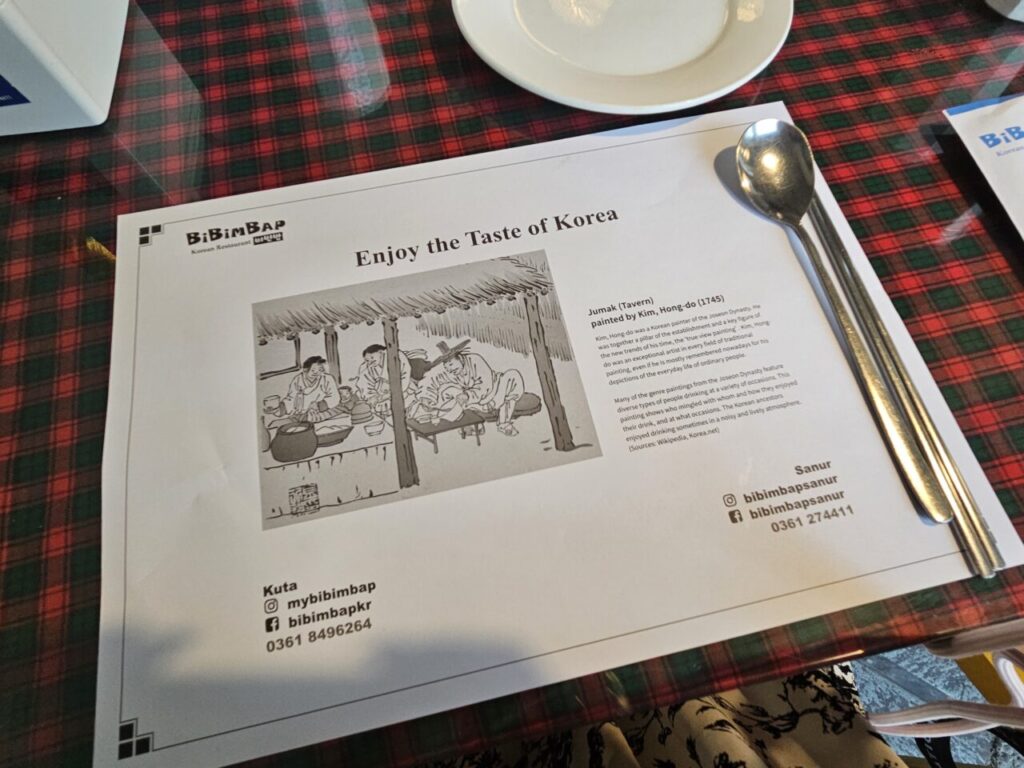체크무늬 테이블보 위에 놓인 발리 비빔밥 한식당의 '한국의 맛을 즐기세요' 문구가 적힌 메뉴/플레이스 매트. 전통 주막 그림과 그에 대한 설명이 있고, 스테인리스 스푼이 함께 올려져 있다.