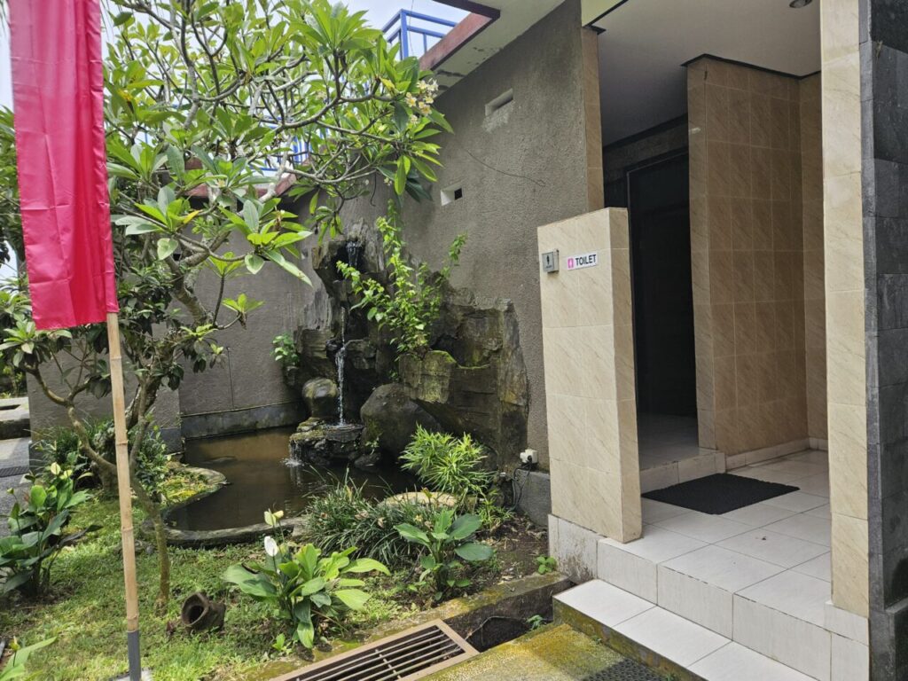 발리 여행 중 발견한 야외 화장실 입구와 그 옆에 아름답게 꾸며진 작은 정원의 모습. 인공 폭포가 있는 연못과 다양한 식물들이 어우러져 이국적인 분위기를 자아낸다. 화장실 표지판과 붉은색 깃발도 보인다.