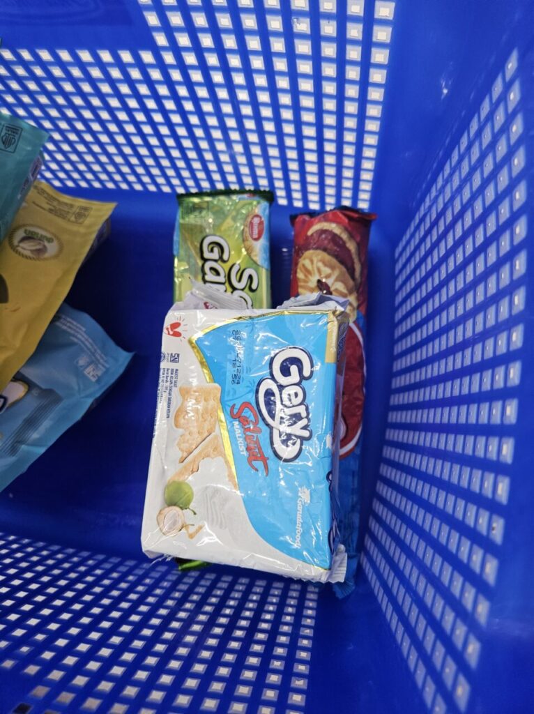 파란색 바구니에 담긴 발리 현지 과자, Gery Saluut Malkist 코코넛 크래커와 다른 스낵 봉투들이 보인다.
