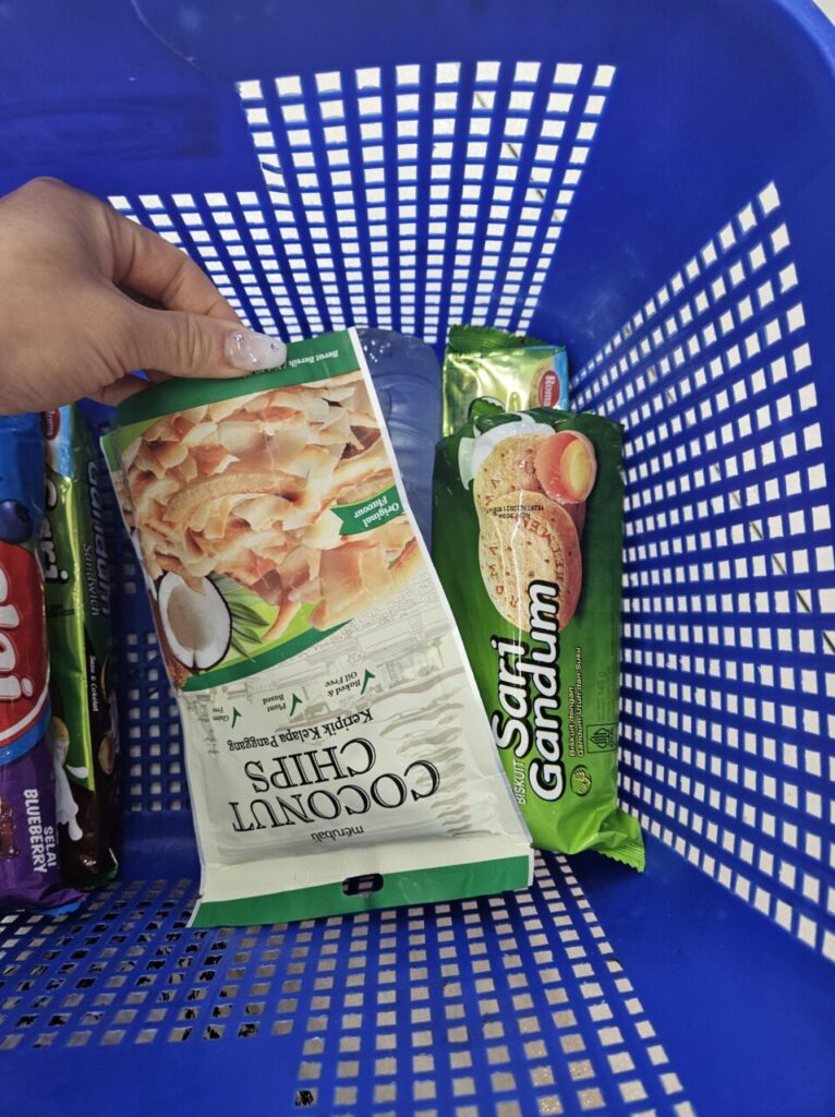 발리 슈퍼마켓에서 파란색 쇼핑 바구니에 담긴 코코넛 칩, 사리 간둠 비스킷 등 현지 간식들. 손으로 코코넛 칩 봉지를 들고 있는 모습.