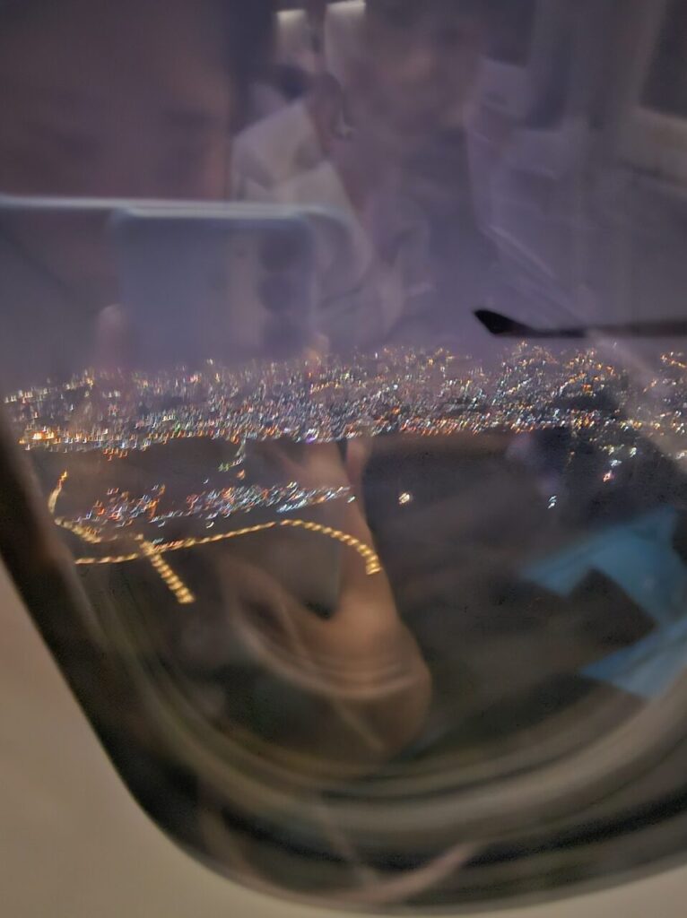 발리 비행 중 창문으로 본 밤 도시의 화려한 불빛 야경과 비행기 날개, 그리고 창문에 비친 기내 승객들의 모습과 스마트폰