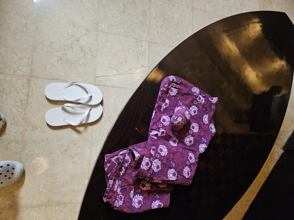 대리석 바닥에 놓인 서핑보드 모양의 어두운 나무 테이블 위에 흰색 슬리퍼 두 켤레와 보라색 패턴의 옷이 놓여 있는 모습.