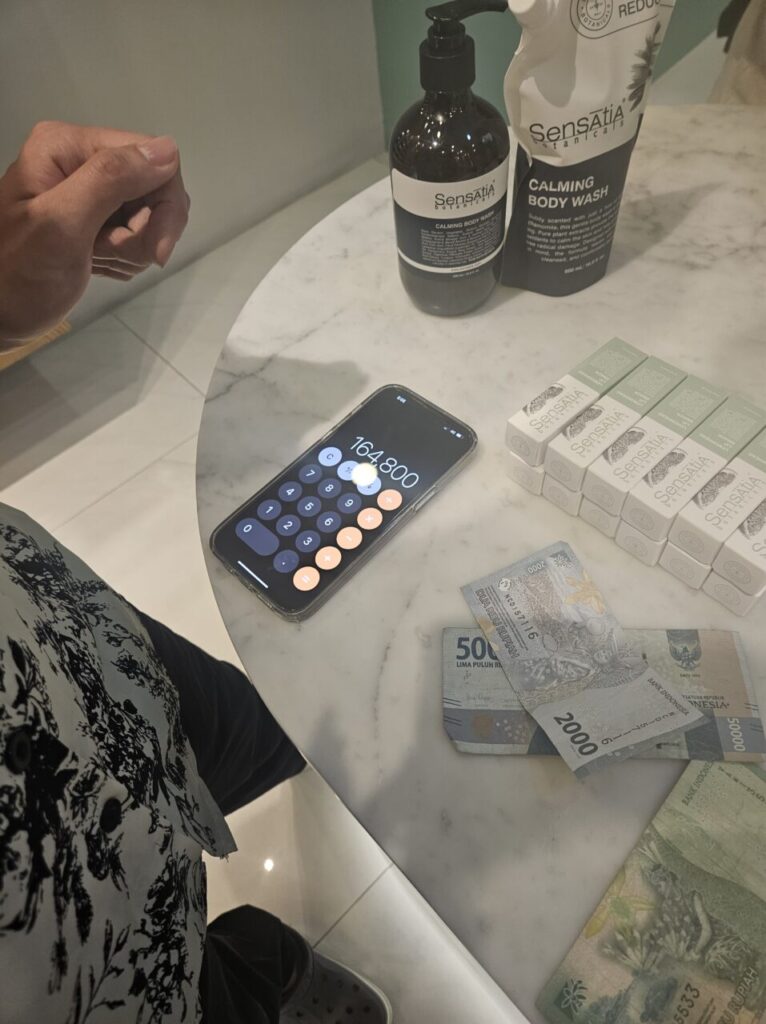 발리 센사티아 보타니컬즈 매장에서 바디워시와 비누를 구매 후 스마트폰 계산기에 164,800 루피아가 표시되고 인도네시아 루피아 지폐가 놓인 쇼핑 계산 장면.