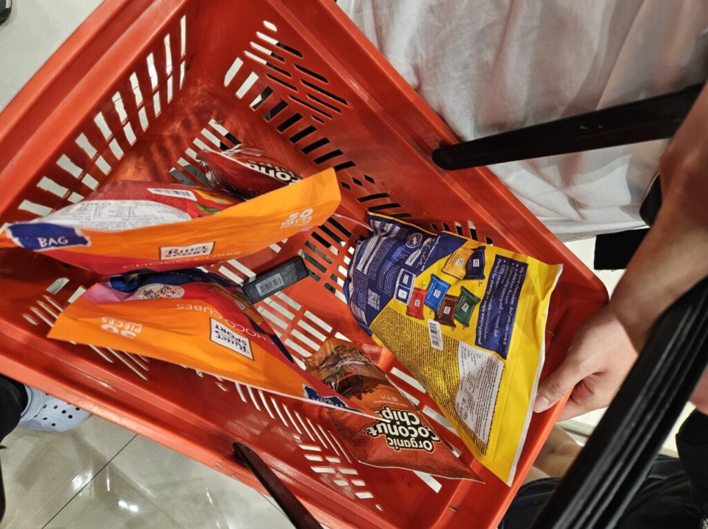 발리 여행 중 빨간 쇼핑 바구니에 리터 스포트 초코 큐브, 코코넛 칩 등 다양한 간식들이 가득 담겨 있는 모습