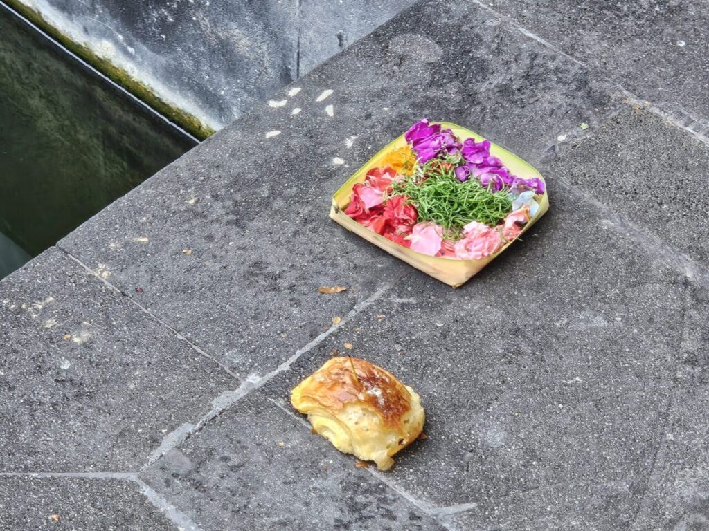 돌바닥 위에 놓인 발리의 전통 제물인 차낭 사리입니다. 작은 바구니에 다채로운 꽃과 풀이 담겨 있으며, 그 옆에는 작은 페이스트리가 함께 놓여 있습니다.