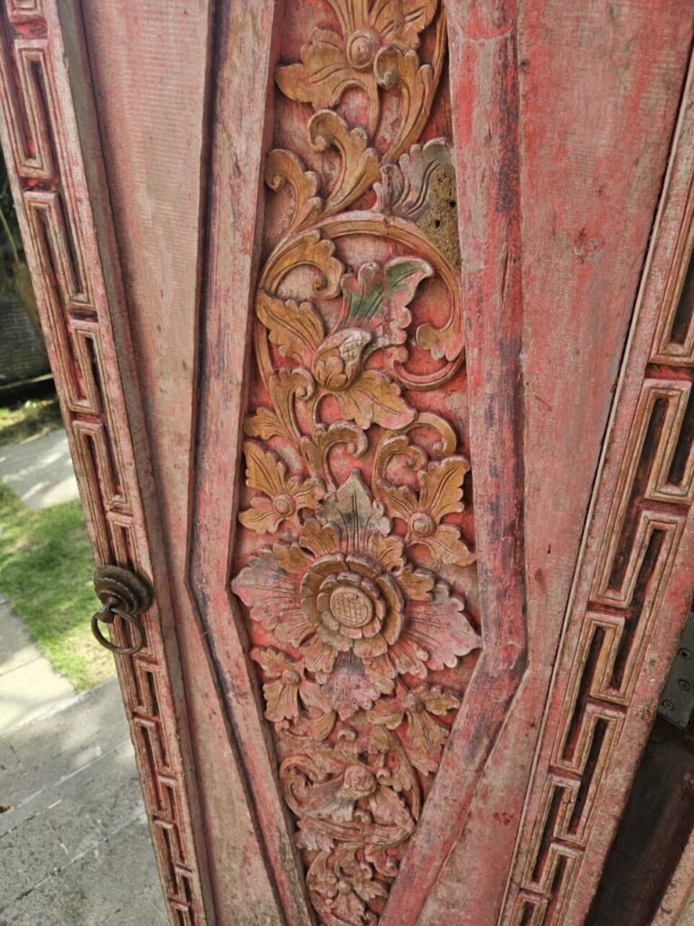 섬세하게 조각된 발리 전통 문양의 붉고 금색 톤으로 칠해진 나무문입니다. 복잡하고 아름다운 꽃과 잎사귀 문양이 돋보입니다.