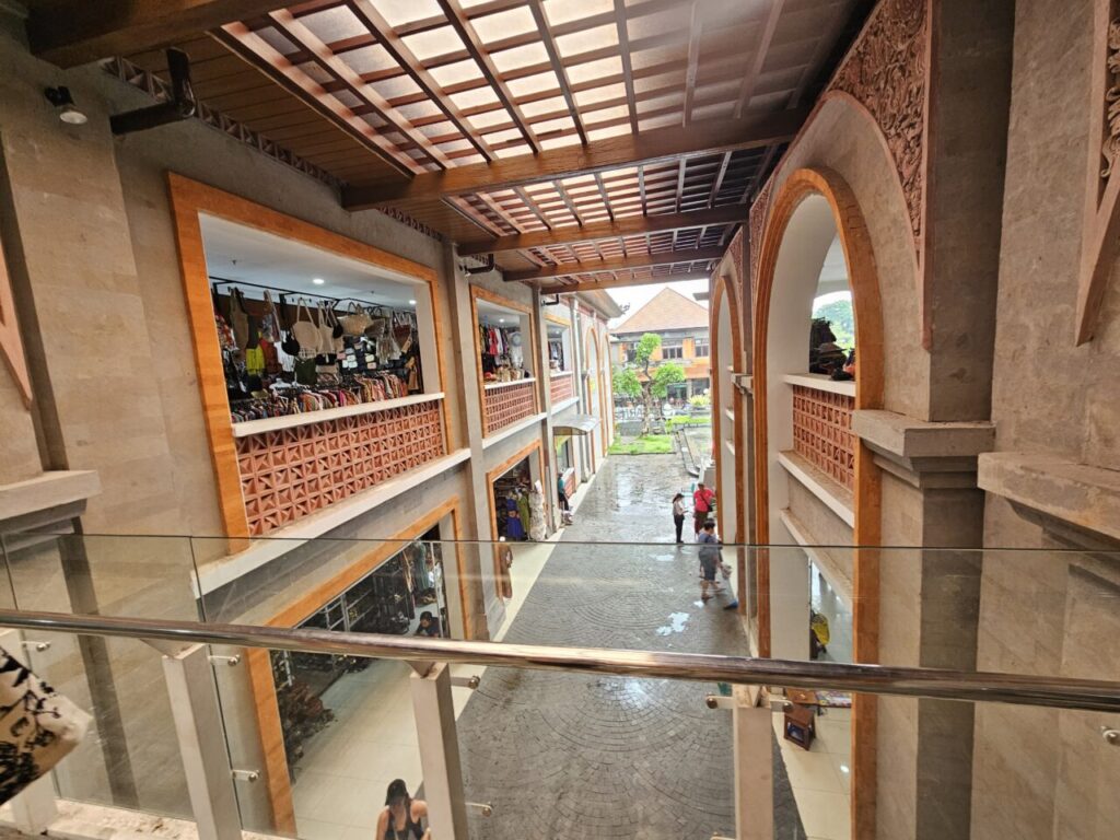 발리 우붓 시장의 다층 쇼핑 공간 전경. 수공예 가방과 의류 등을 판매하는 상점들이 층층이 보이며, 발리 전통 양식의 건축물과 개방형 천장이 특징이다. 중앙 야외 공간에는 사람들이 이동하고 있다.