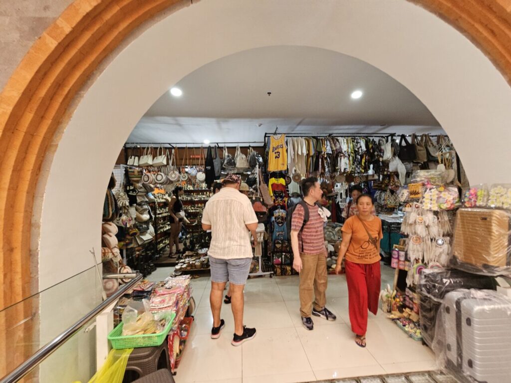 아치형 입구 너머로 발리 우붓 시장의 활기찬 내부 풍경이 보인다. 다양한 디자인의 가방, 옷, 기념품, 캐리어 등을 구경하는 관광객들의 모습.