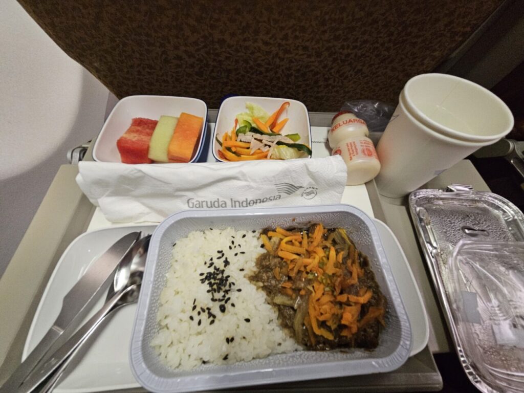 가루다 인도네시아 항공 기내식 트레이에 쌀밥, 고기 볶음, 과일(수박, 멜론, 칸탈루프), 샐러드, 야쿠르트가 정갈하게 놓여 있다.