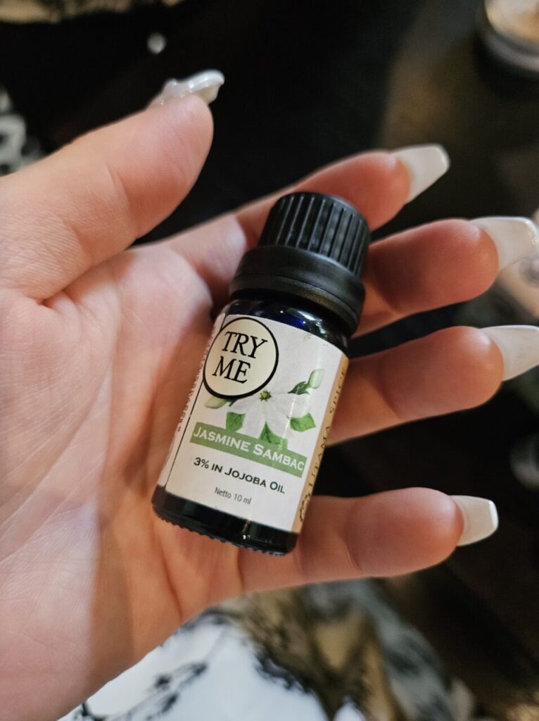 'JASMINE SAMBAC 3% IN JOJOBA OIL'이라고 쓰인 작은 파란색 자스민 삼박 오일 병을 들고 있는 손.