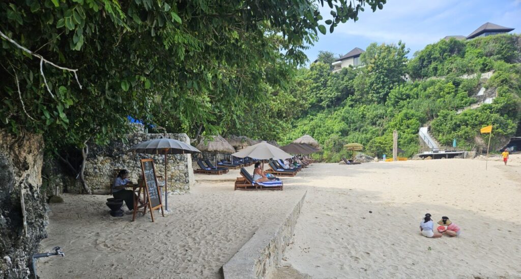 발리 쿠부 비치의 한적한 모래 해변. 나무 그늘 아래 선베드와 파라솔이 놓여 있고, 사람들이 휴식을 취하고 있다.