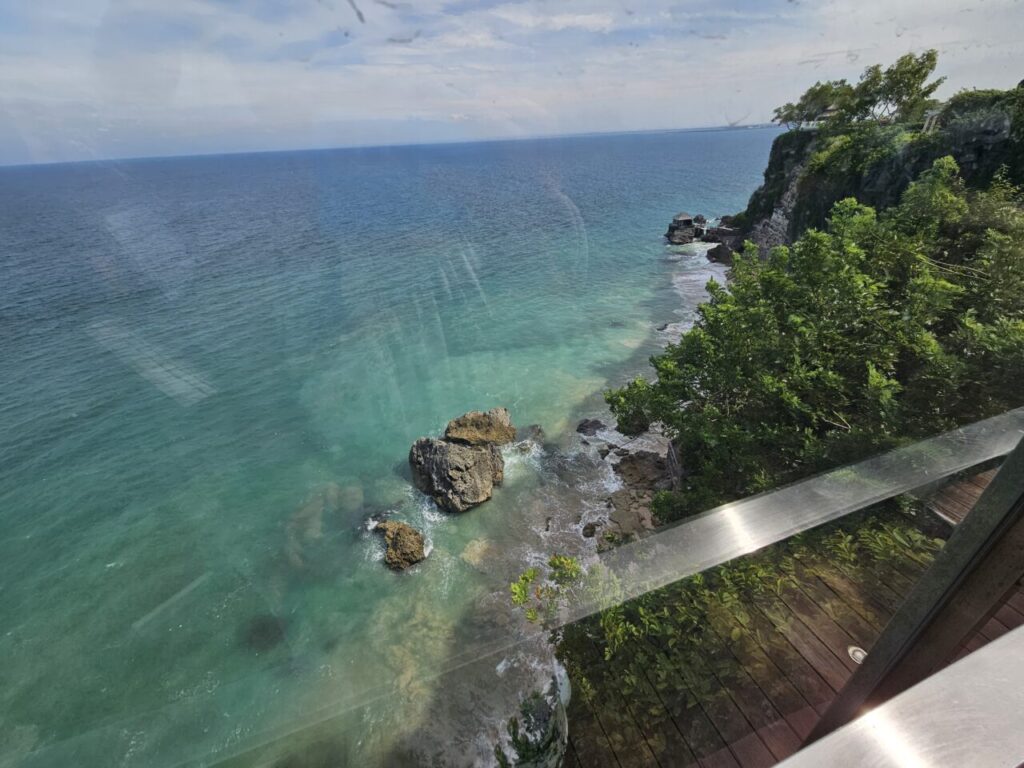 발리 쿠부 비치 절벽에 설치된 유리 엘리베이터 안에서 바라본 푸른 바다와 해안선 풍경.