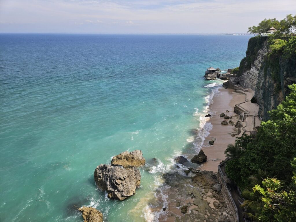 발리 쿠부 비치의 절벽 위에서 바라본 에메랄드빛 바다와 해변 전경. 큰 바위들이 해안선을 따라 펼쳐져 있다.