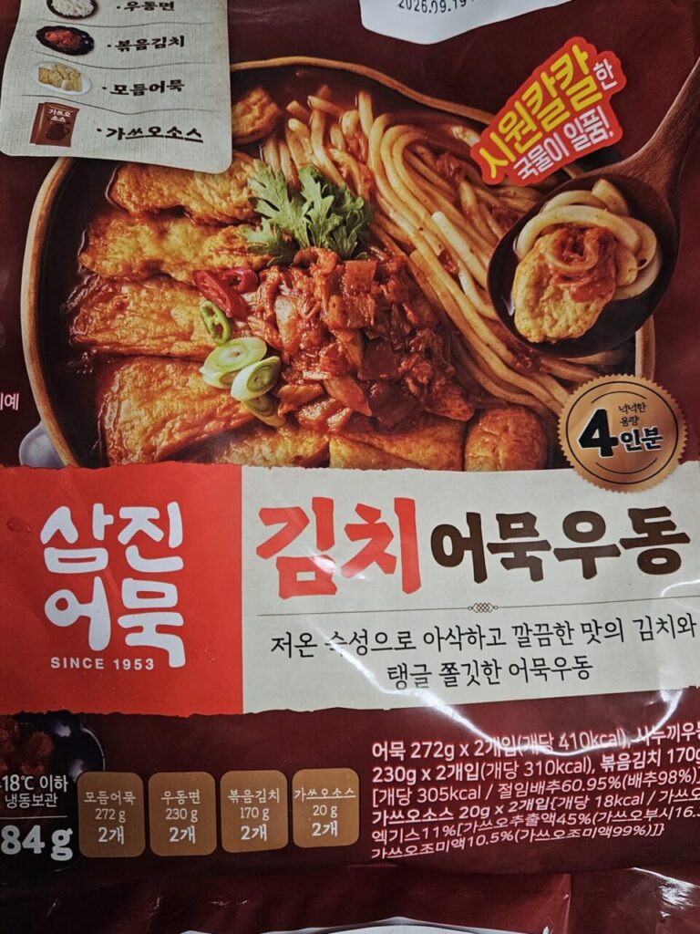 '삼진어묵 김치어묵우동' 제품 포장지입니다. 김치, 어묵, 우동 면이 어우러진 요리의 이미지가 있고, 4인분이라고 명시되어 있습니다. 시원칼칼한 국물이 특징입니다.