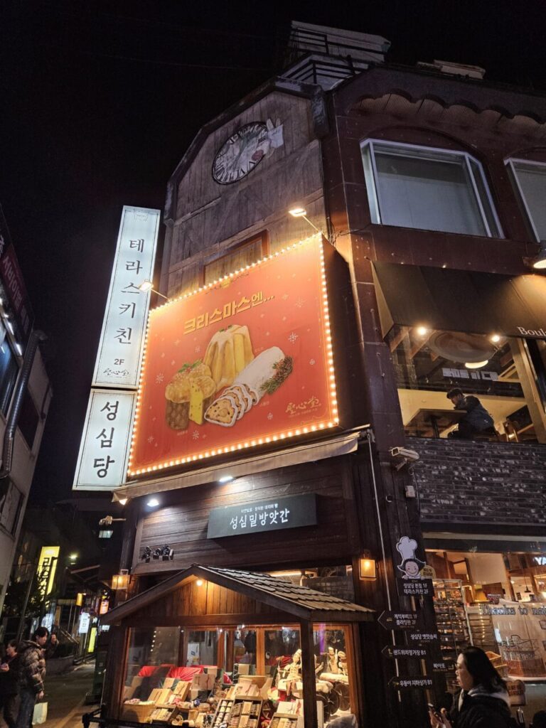 밤에 촬영된 성심당 건물 외관으로, 커다란 크리스마스 포스터가 밝게 빛나고 있습니다. '테라스 키친', '성심당 옛맛솜씨' 등 여러 상점의 간판이 함께 보입니다.