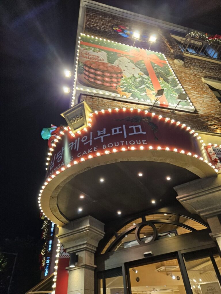 밤에 조명으로 환하게 빛나는 성심당 케익 부띠끄 외관입니다. 건물 상단에는 커다란 크리스마스 테마의 광고판이 걸려 있고, 입구 주변은 전구로 장식되어 있습니다.