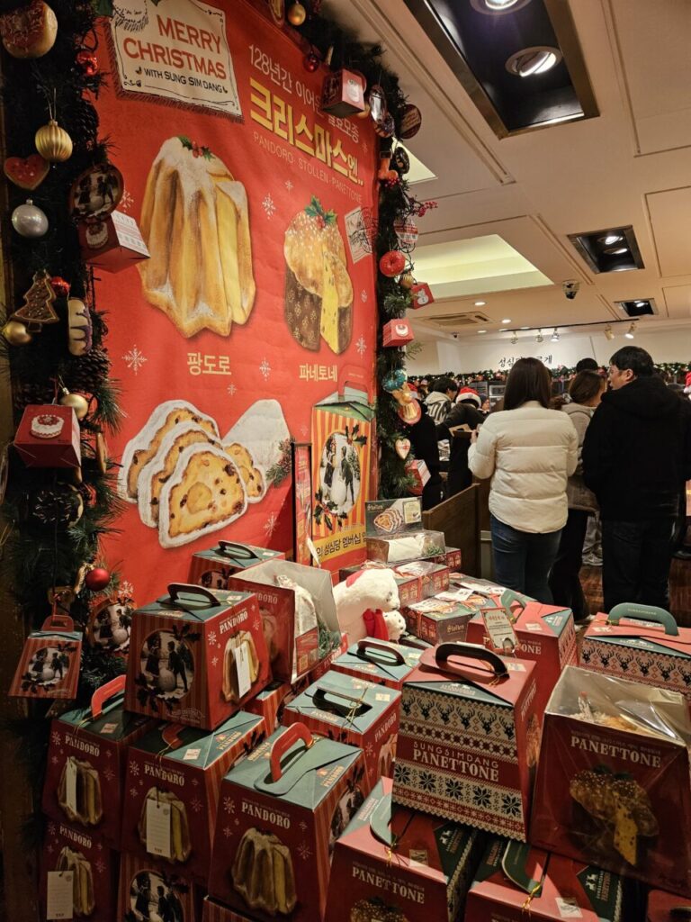 성심당 베이커리에서 크리스마스 시즌을 맞아 판도로와 파네토네를 홍보하는 대형 빨간색 배너가 걸려 있습니다. 그 아래에는 크리스마스 디자인의 선물 상자들이 쌓여 있습니다.