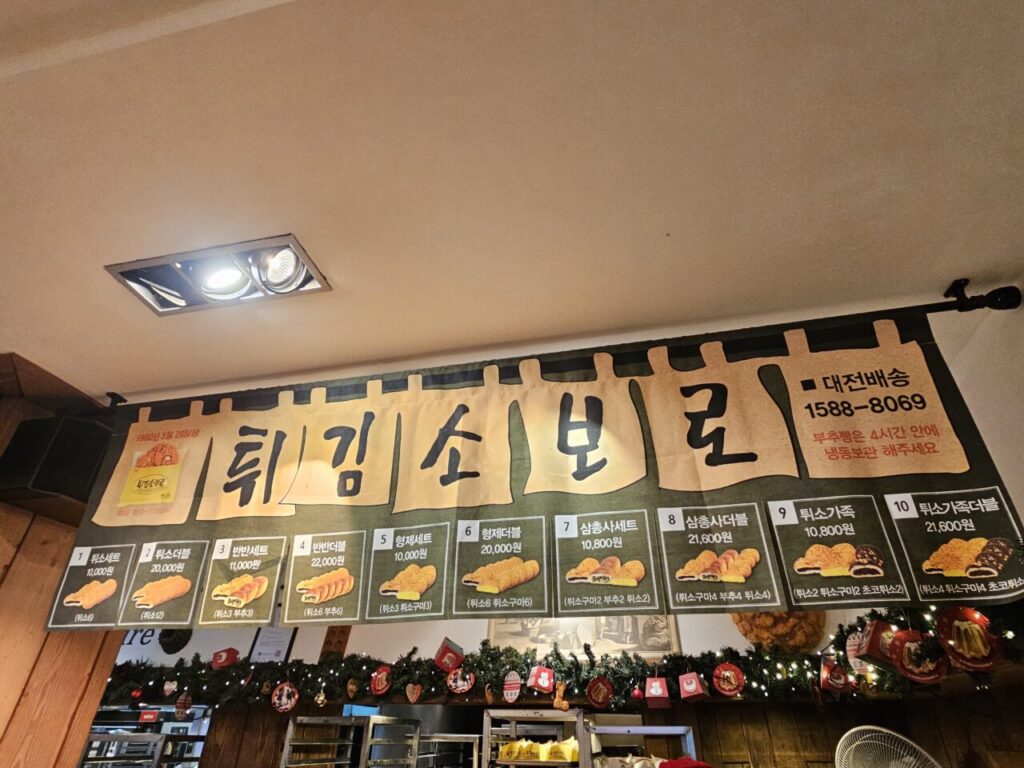 성심당의 시그니처 메뉴인 '튀김소보로' 세트 상품들을 소개하는 대형 천장 배너입니다. 다양한 튀소 세트 구성과 가격, 그리고 대전 배송 서비스 안내가 포함되어 있습니다.