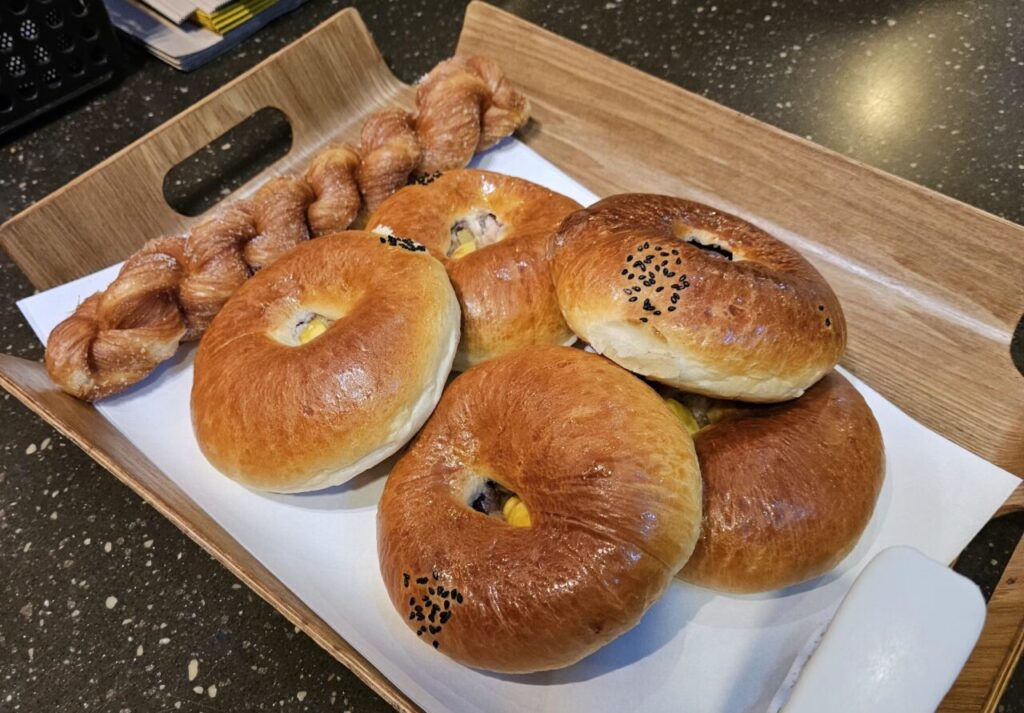 나무 트레이에 담긴 여러 개의 튀소구마와 튀소구마 도넛처럼 보이는 둥글고 노릇한 빵들, 그리고 설탕이 뿌려진 꽈배기 형태의 페이스트리입니다.