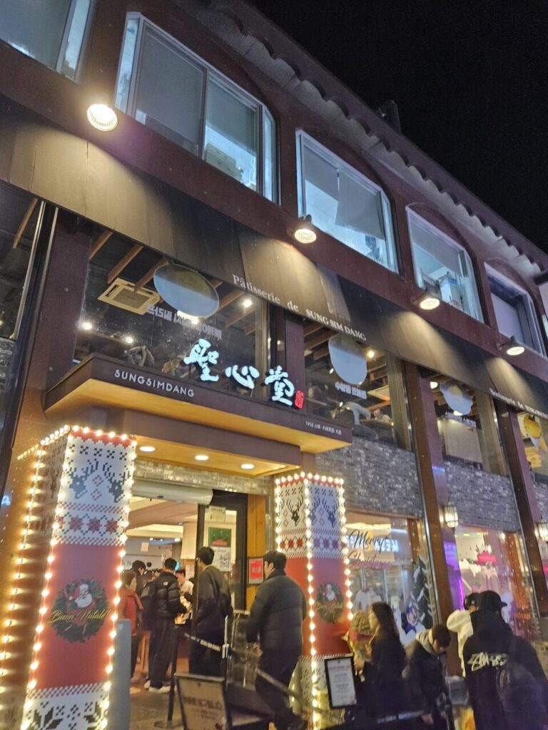 밤에 화려한 조명으로 장식된 성심당 건물의 외관입니다. 'Patisserie de SUNGSIMDANG'과 한자 '聖心堂' 간판이 빛나고 있으며, 많은 사람들이 출입하는 모습이 보입니다.