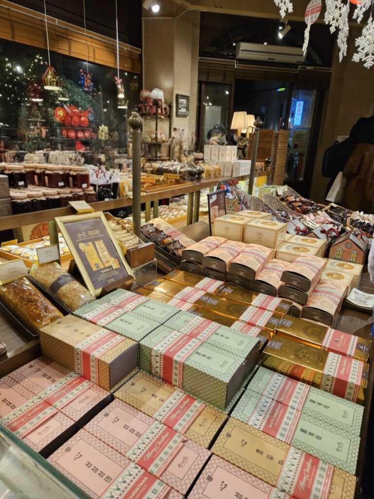 대전 성심당 스트리트의 크리스마스 분위기로 장식된 베이커리 내부 전경입니다. 다양한 빵과 크리스마스 선물 세트들이 진열대에 가득하며, 손님들이 제품을 구경하고 있습니다.