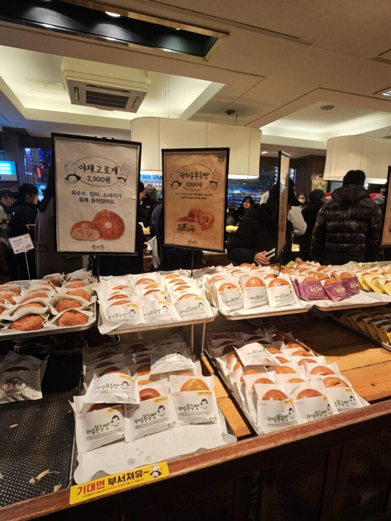 성심당 베이커리 카운터에 개별 포장된 '야채고로케'와 '판타롱부추빵'이 가득 진열되어 있습니다. 각 빵의 가격과 설명이 적힌 포스터가 함께 보입니다.